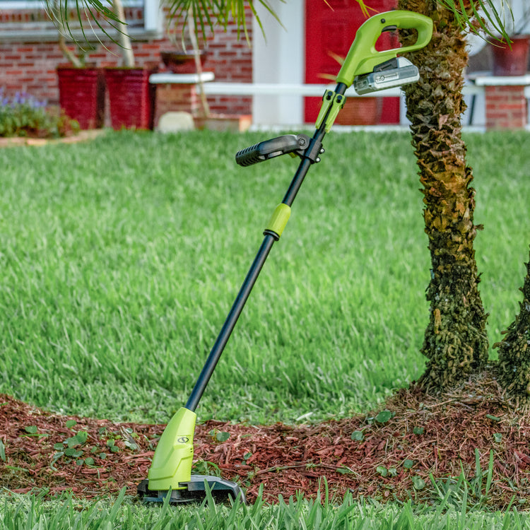 Sun Joe 24-volt cordless 10-inch stringless grass trimmer with a 2.0-Ah lithium-ion battery leaning against a tree.