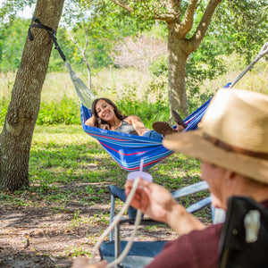 Woman relaxing in the Bliss Hammocks 40-inch Wide America's Cup Hammock in a Bag haning between two trees. She's smiling at a man in the foreground.