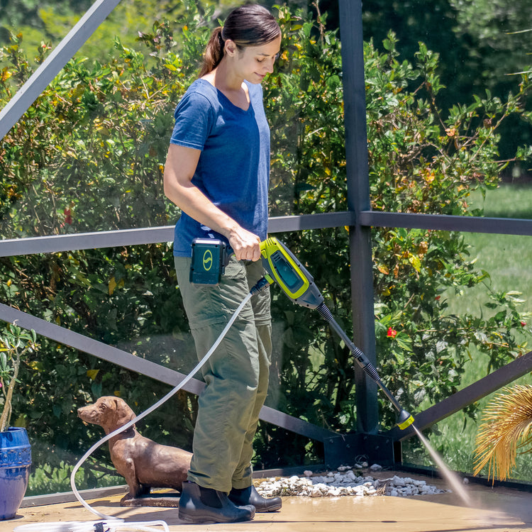 Woman using her Sun Joe 24-Volt Cordless Power Cleaner to clean her patio. Her small dog is behind her.