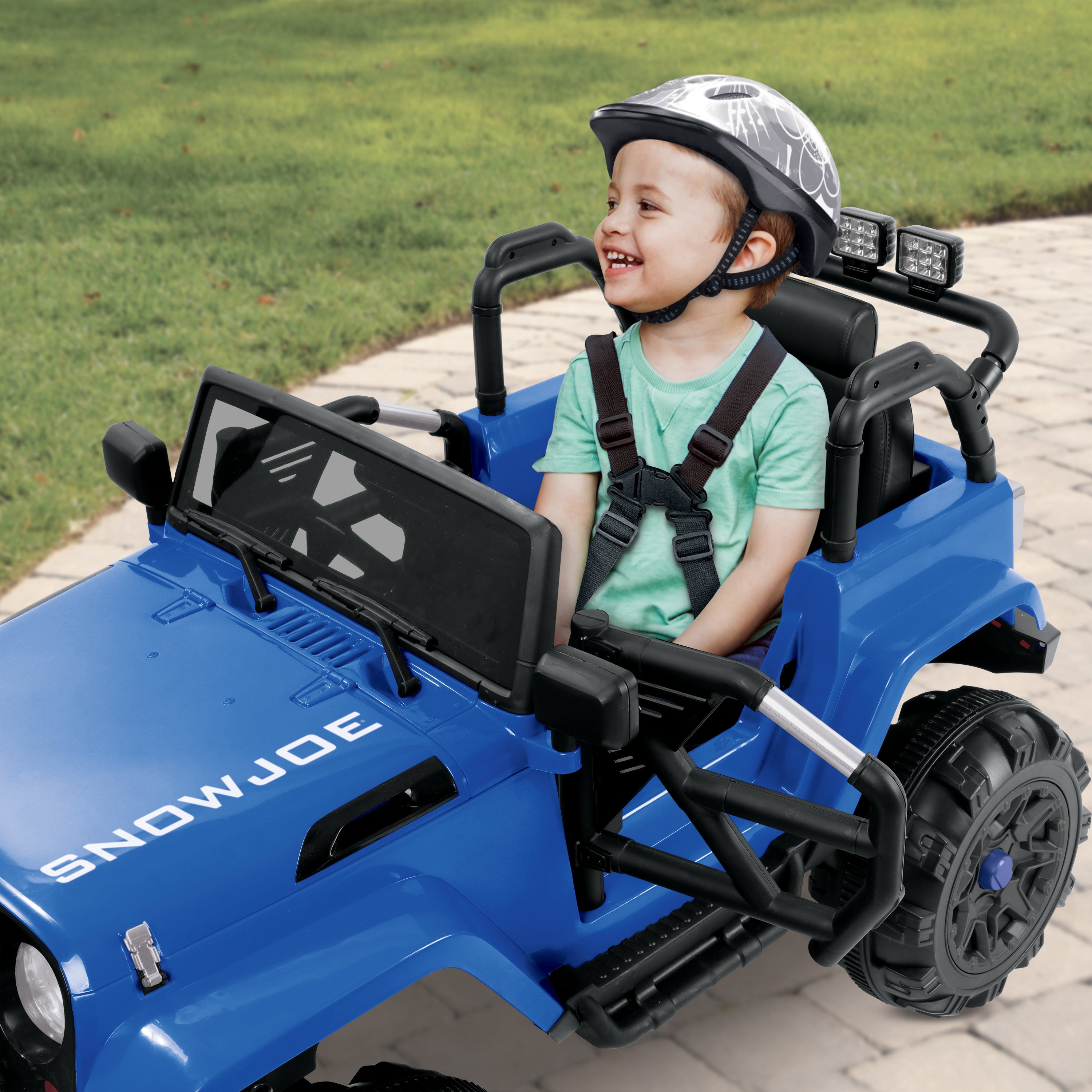 Young boy smiling while wearing a helmet and driving the Snow Joe 24-Volt Ride On Kids Truck in a driveway.