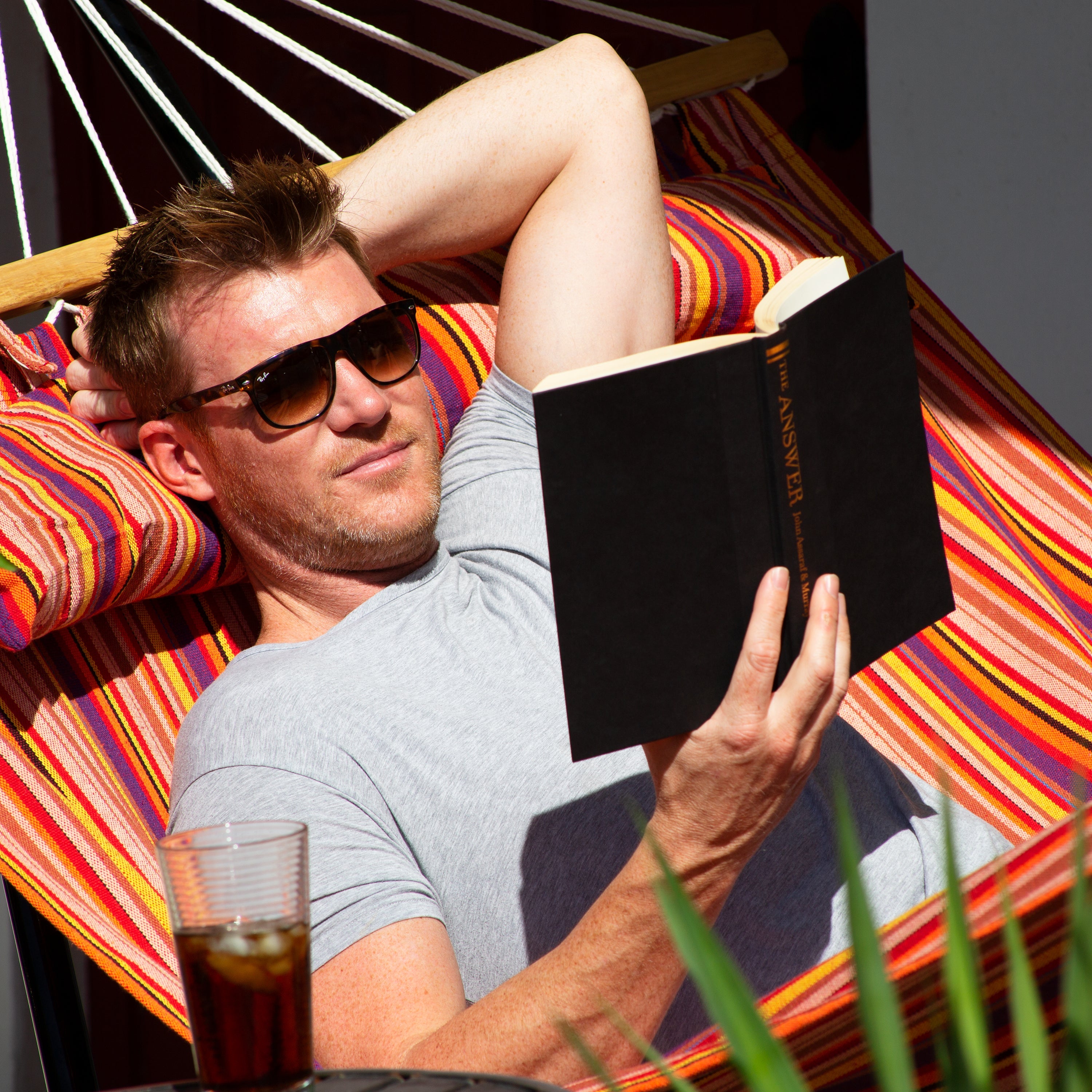 Man reading a book on the Bliss Hammocks 48-inch Wide Toasted Almond Caribbean Hammock with a drink beside him.