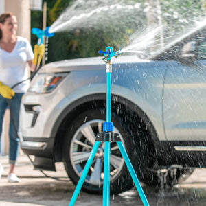 Aqua Joe 72-inch Indestructible Zinc Impulse 360-Degree Telescoping Tripod Sprinkler spraying water while a woman washes a car in the background.