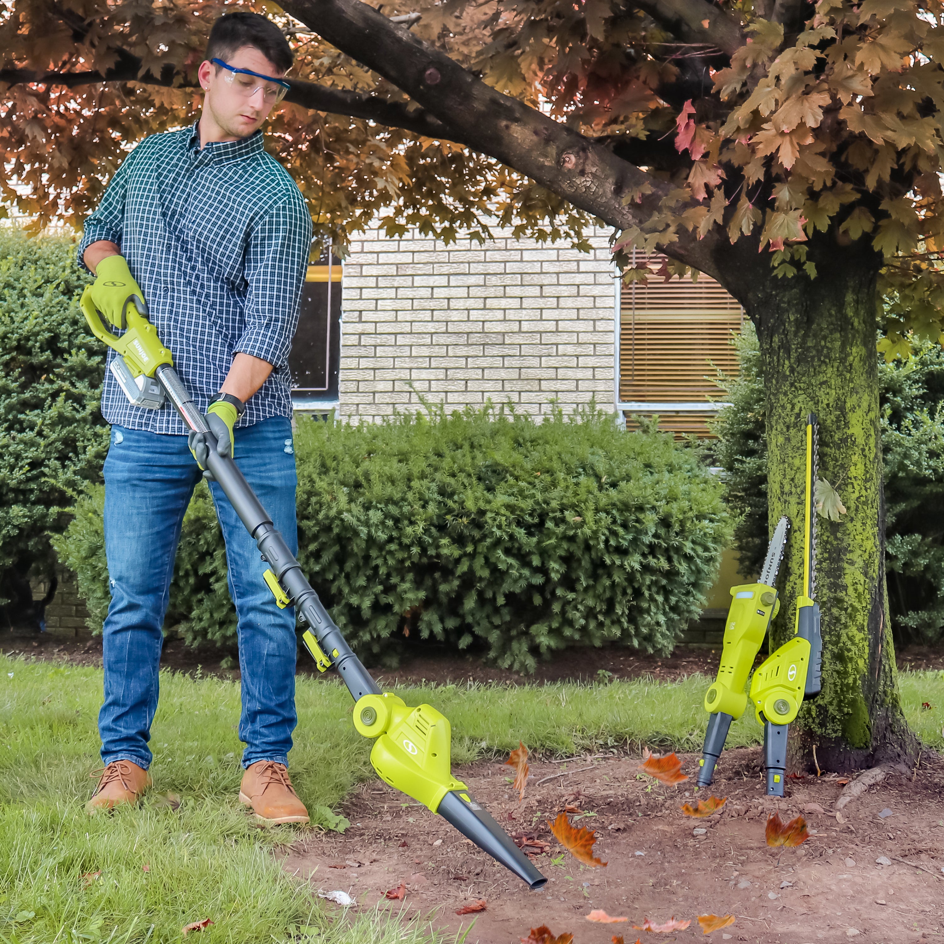 Man using the leaf blower attachment to blow leaves away from the ground around a tree. The pole saw and hedge trimmer attachments are leaning on the tree.