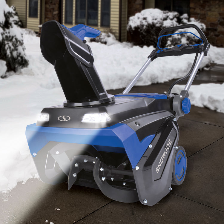 Snow Joe 96-volt cordless 21-inch snow blower outside on a walkway with the LED headlights turned on.